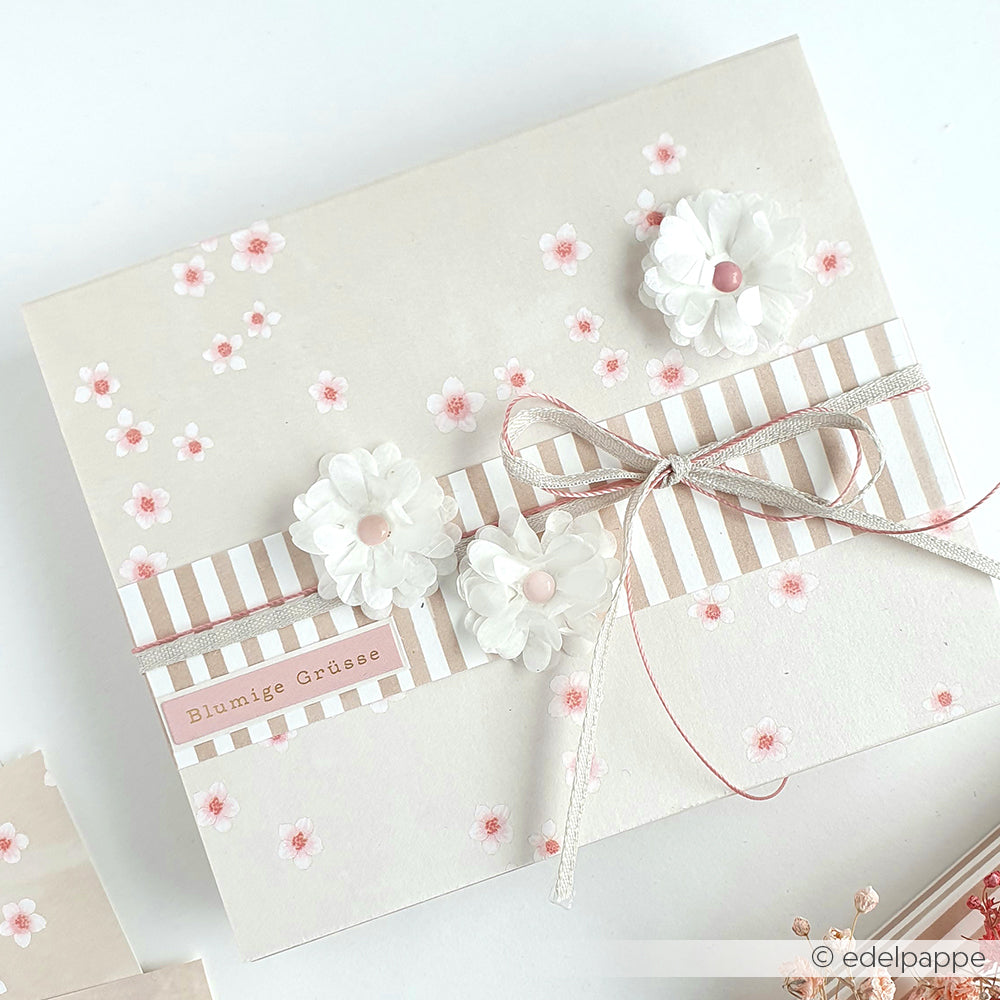 Cremefarbene Karte mit rosa Blüten und weißen Papierblumen aus dem Designpapier Kirschblüten von Alexandra Renke, veredelt mit einer Schleife. Mit dem Text "Blumige Grüße" - ideal für Papierprojekte oder Buchbinderei.