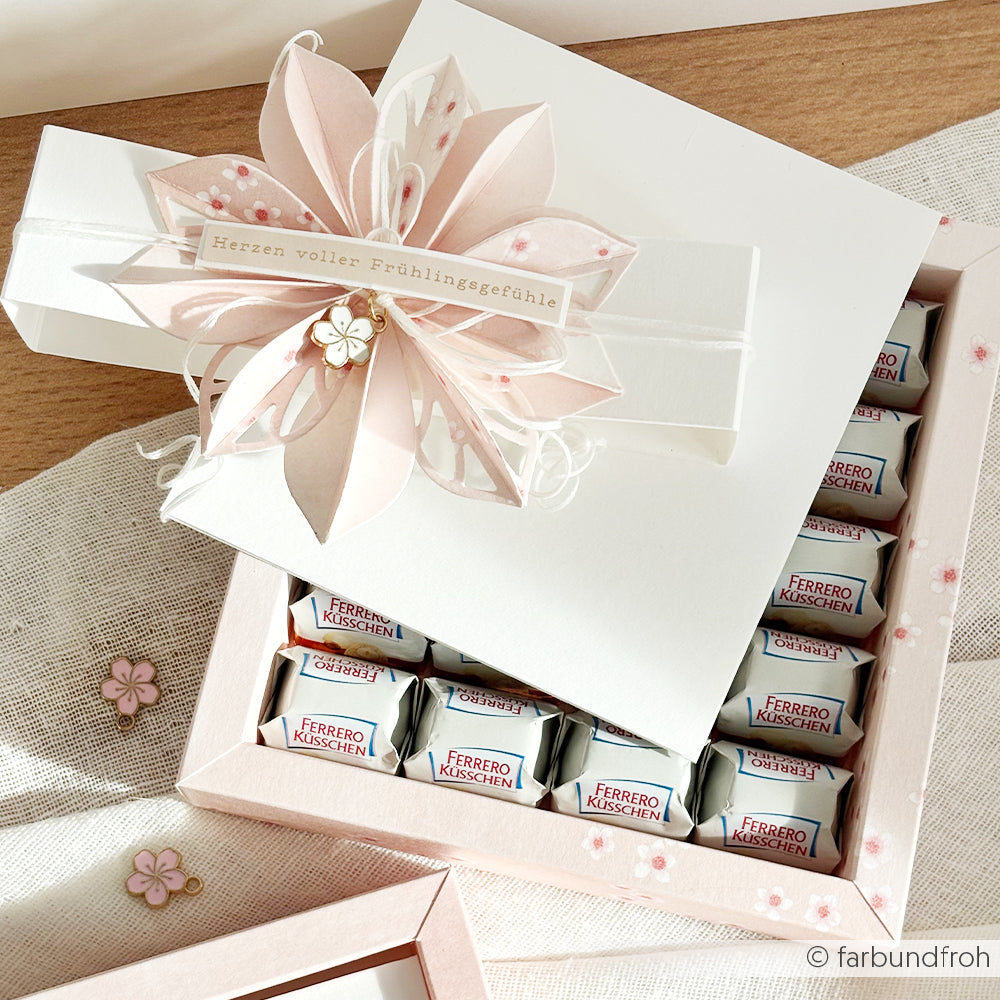 Ein weißer Geschenkkarton von Alexandra Renke mit einem rosafarbenen Kirschblütenmotiv auf elegantem Designpapier, in dem Ferrero Küsschen-Pralinen fein säuberlich angeordnet sind.