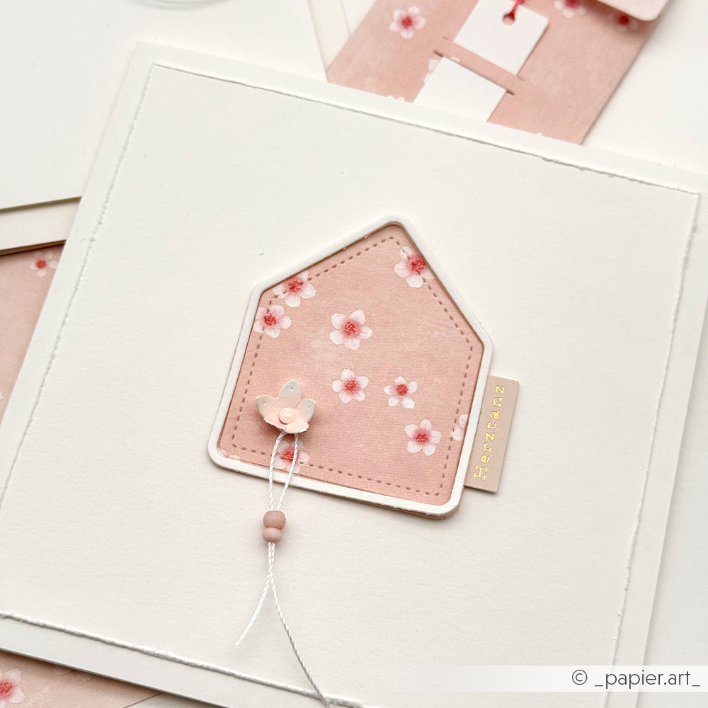 Eine handgefertigte Karte von Alexandra Renke mit einem rosa Blumenhausausschnitt von Designpapier, Kirschblüten-Akzenten, einem kleinen Schmetterling und einer Perlenschnur auf weißem Papier.