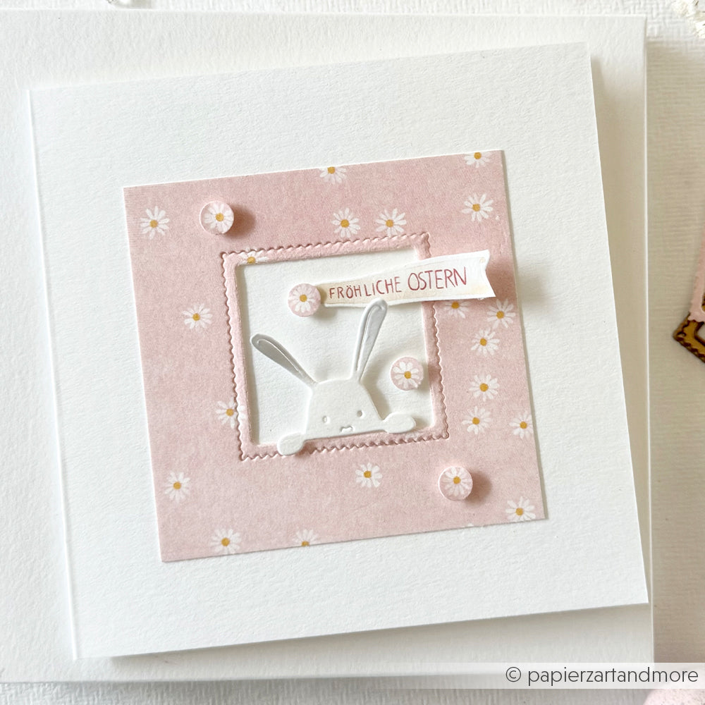 Osterkarte von Alexandra Renke mit einem Hasen, der durch einen rosafarbenen Gänseblümchenrahmen aus Altrosa Designpapier lugt, und einem "Fröhliche Ostern"-Banner.