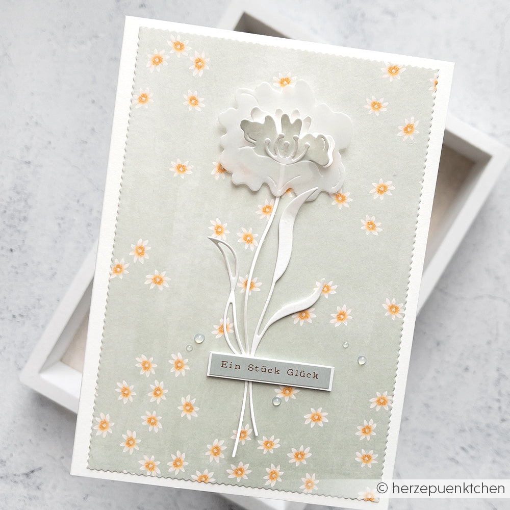 Alexandra Renke-Grußkarte mit weißer Prägeblume und Text "Ein Stück Glück" auf Designpapier vor hellgrünem, floralem Hintergrund - ideal für Papierprojekte oder zur Hervorhebung von Lieblingsblüten auf Greige.