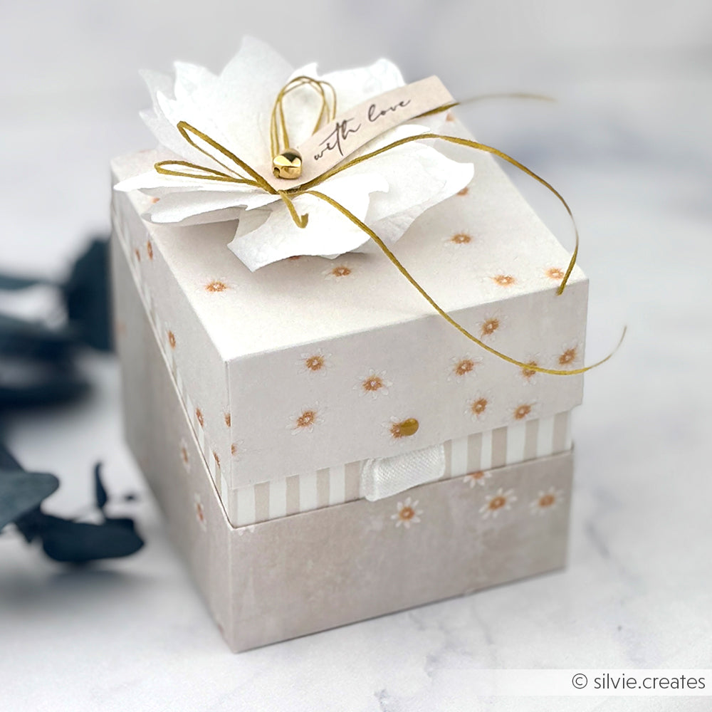 Quadratische Blumengeschenkbox von Alexandra Renke mit einer weißen Papierblume aus Designpapier, die mit goldener Schnur gebunden und mit "with love" beschriftet ist und auf einer Marmoroberfläche präsentiert wird.