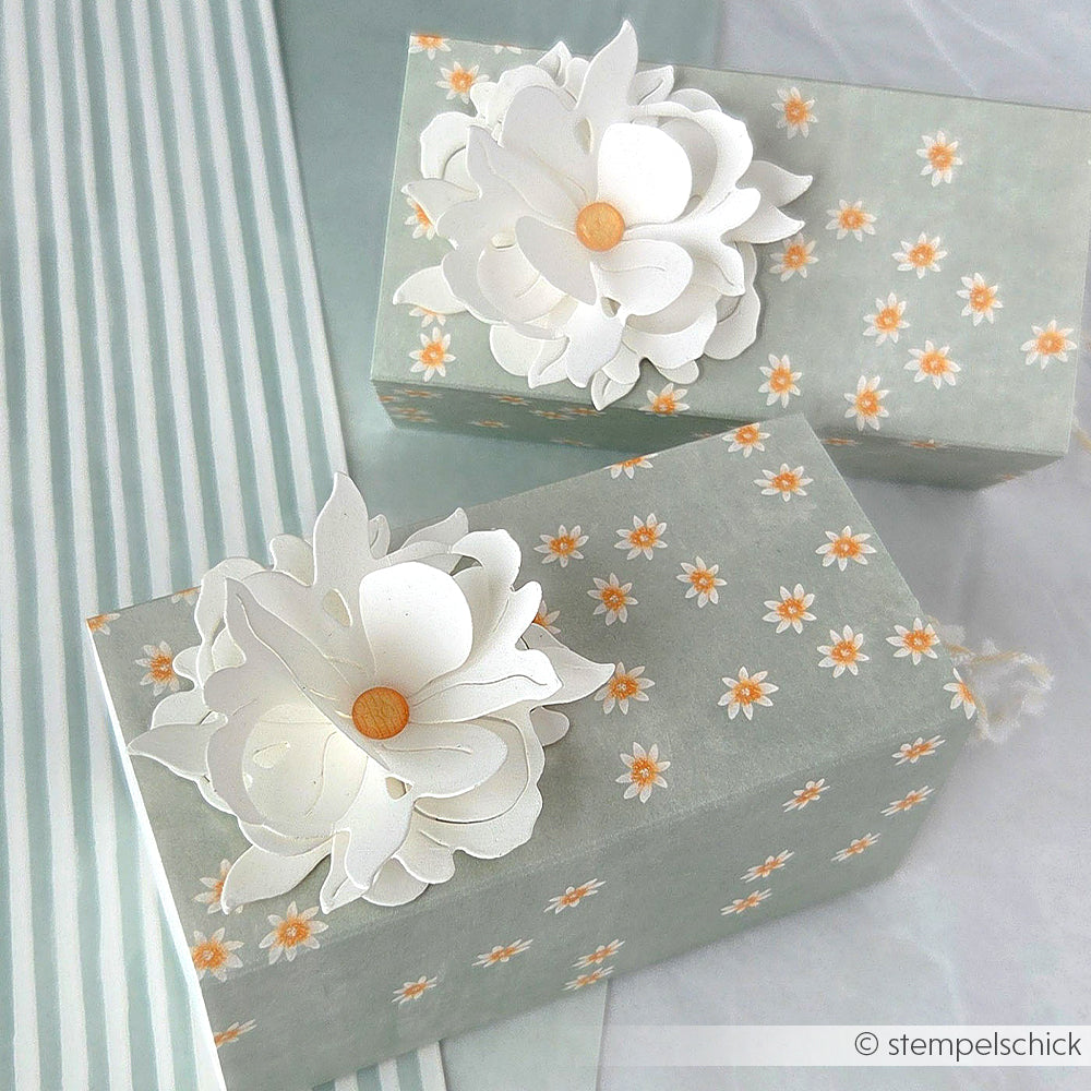 Zwei Geschenkboxen mit floralen Mustern und großen weißen Papierblumen auf der Oberseite, gefertigt aus Alexandra Renke Designpapier in zartem Sagegrün, stehen auf einer gestreiften Fläche.