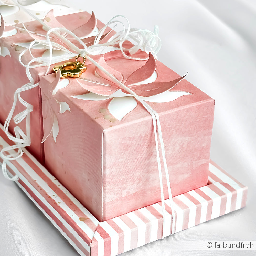 Geschenkschachteln in rosa und weiß, hergestellt aus Alexandra Renke Designpapier Korallrosa, verziert mit Blumen und einem goldenen Vogelanhänger, veredelt mit weißem Band - ideal für Papierprojekte oder Buchbinderei.