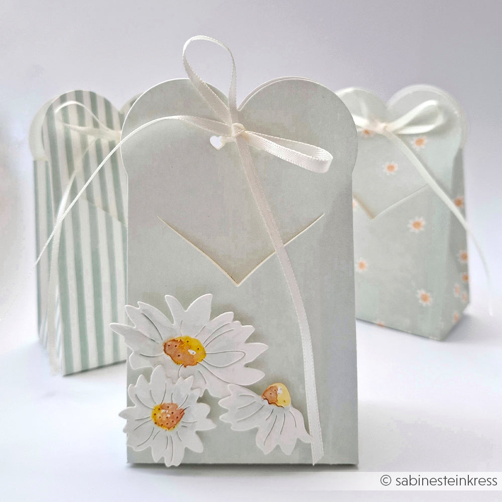 Drei pastellfarbene Geschenktüten, gefertigt mit Alexandra Renke Designpapier Calm Jadeblau und verziert mit Schleifen und weißen Gänseblümchen, stehen auf einer weißen Fläche - ideal für Papierprojekte oder zum stilvollen Überreichen von Grußkarten.
