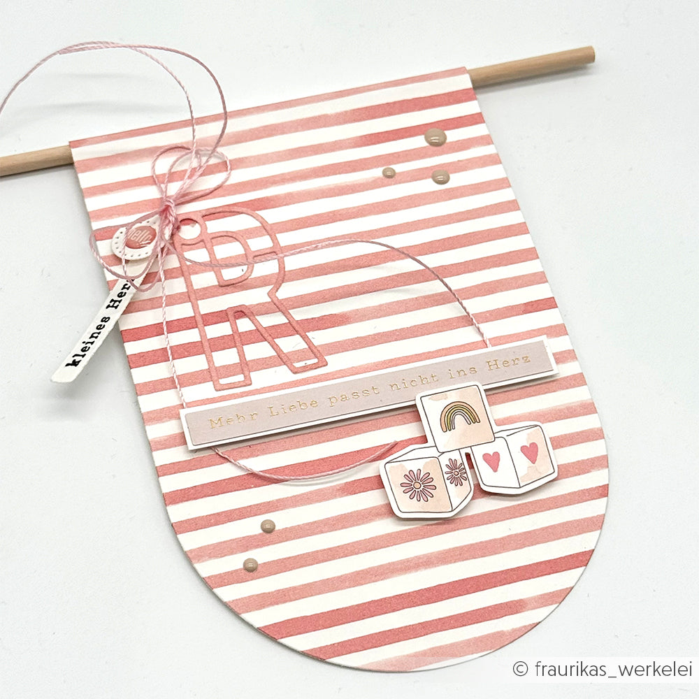 Ein rosa-weiß gestreifter Wandbehang aus Alexandra Renke Designpapier Streifen Korallrosa, mit dem Buchstaben "R", Herzblöcken und dem Text "Mehr Liebe passt nicht ins Herz". Ideal für Buchbinderei oder Papierprojekte!.
