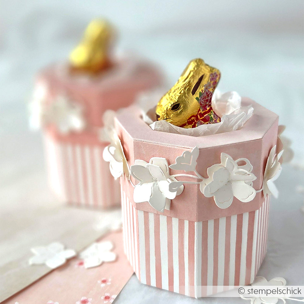 Zwei rosa-weiße Geschenkboxen von Alexandra Renke aus Designpapier mit Papierblumen und goldenen Schokoladenhasen im Inneren.
