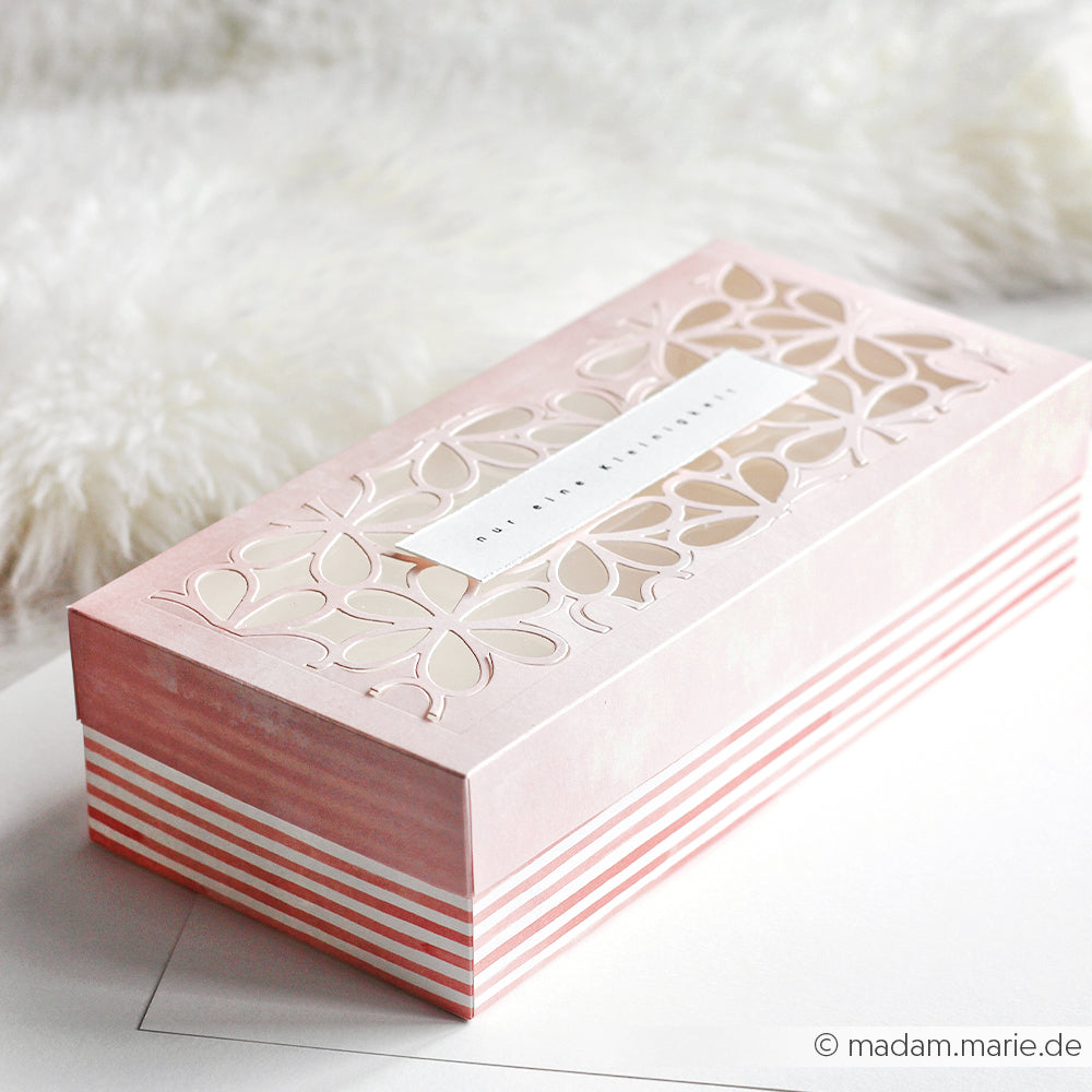 Rosa Dekobox von Alexandra Renke mit floralen Ausschnitten und gestreiften Korallrosa-Seiten, ideal für Papierprojekte oder Buchbinderei, ausgestellt auf einer weißen Fläche neben weichem Stoff.