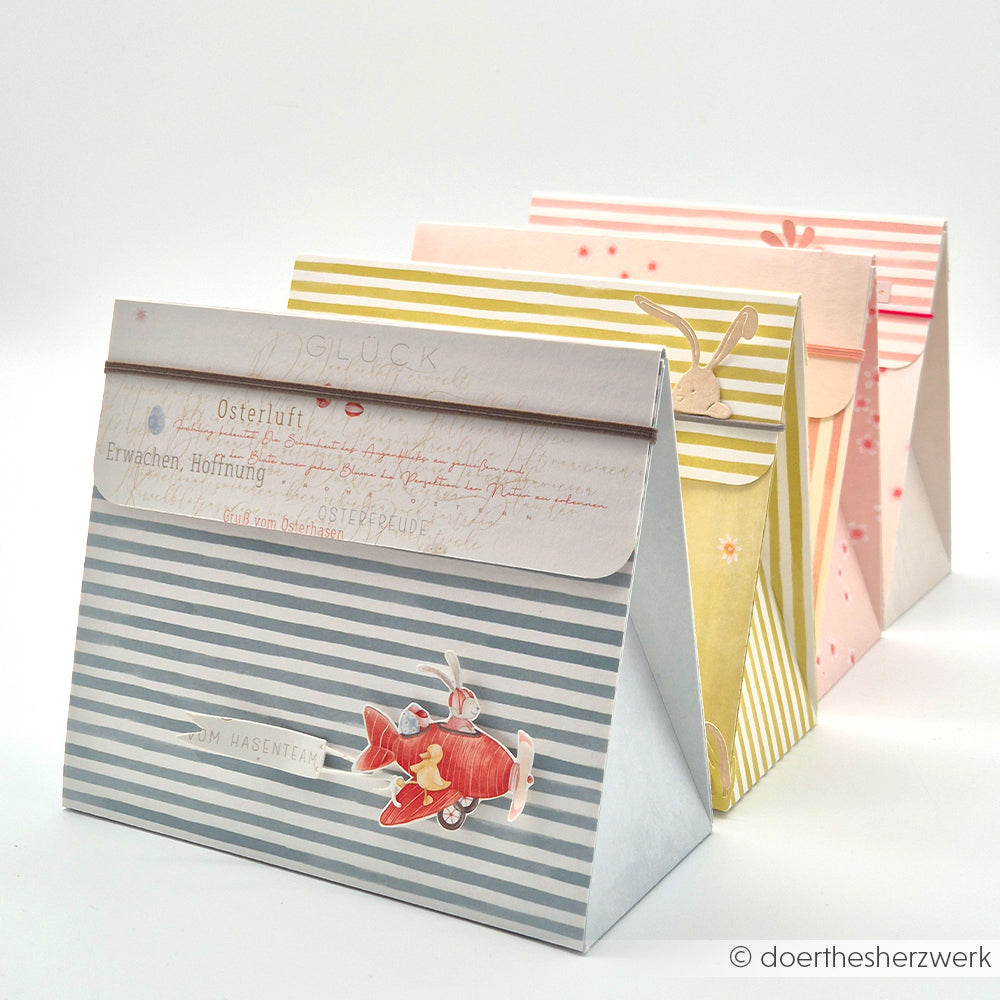 Drei pastellfarbene Geschenktüten von Alexandra Renke aus Designpapier in Jadeblau und zarten Tönen mit Hasen- und Flugzeugmotiven, arrangiert auf weißem Hintergrund - ideal für kreative Papierprojekte oder zum Verschenken.
