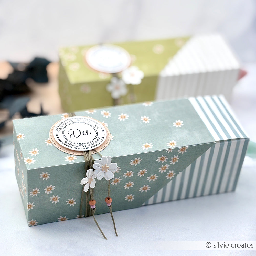 Dekorative florale Geschenkschachtel von Alexandra Renke mit einem Du-Tag, Blumenverzierungen und Designpapier Streifen Jadeblau - ideal für Papierprojekte oder zum Verschenken mit Grußkarten. Im Hintergrund ist eine weitere passende Schachtel zu sehen.