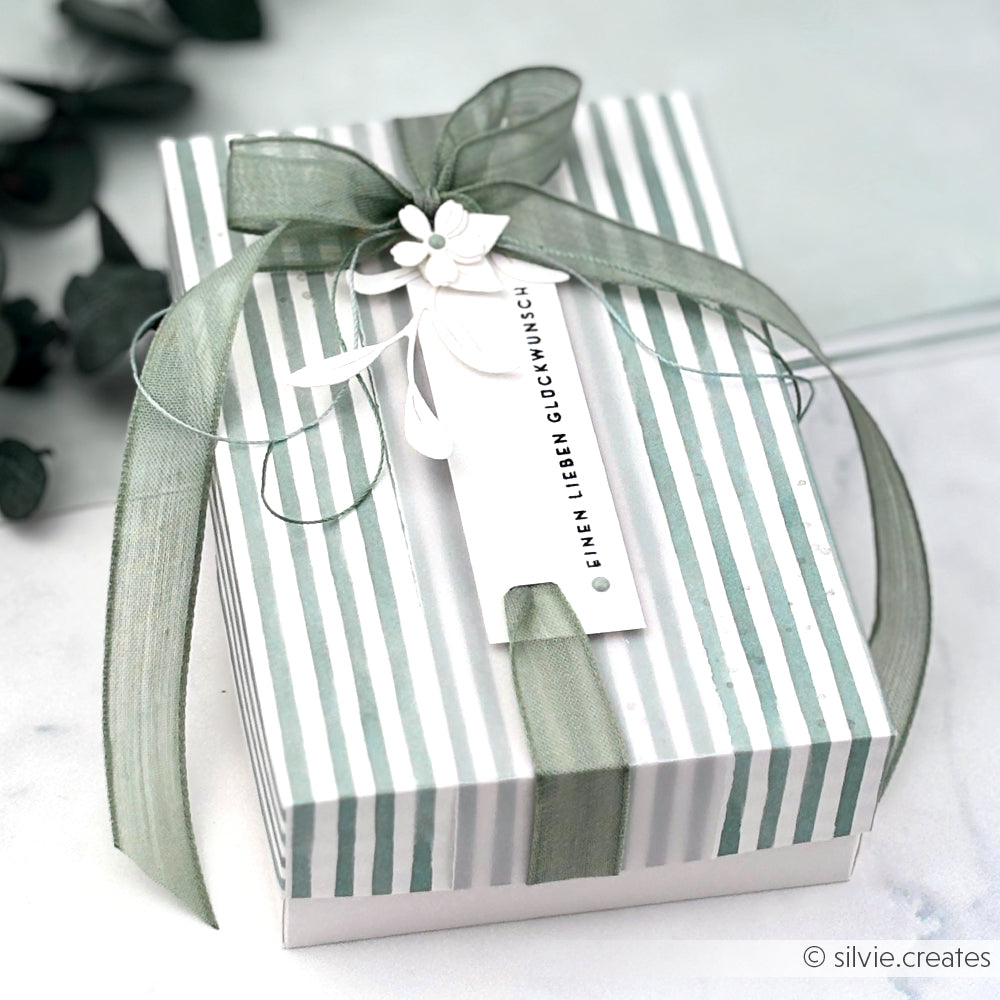 Gestreifte Geschenkbox mit grünem Band, Anhänger und kleiner weißer Blumendekoration - ideal für Papierprojekte oder Buchbinderei mit Alexandra Renke Designpapier Streifen Jadeblau.
