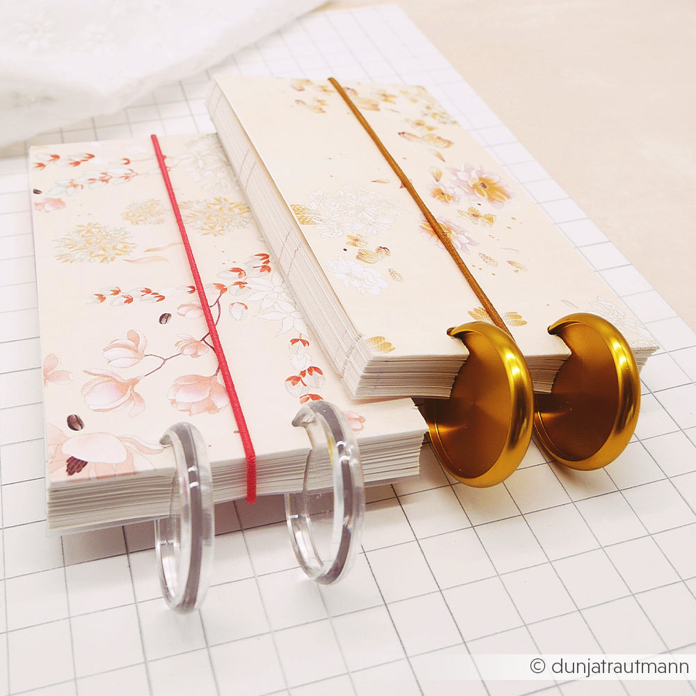 Zwei floral gemusterte Alexandra Renke Notizbücher mit dekorativer Ringbindung, gesetzt auf gerastertem Papier - ideal für Papierprojekte oder zur Darstellung einer floralen Blütenkomposition auf hochwertigem Designpapier.