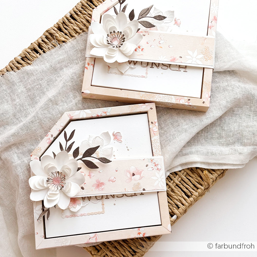 Zwei mit Blumenmotiven verzierte Geschenkboxen von Alexandra Renke, die eine zarte florale Blütenkomposition mit zartrosa und weißen Details zeigen, stehen elegant auf einem Leinentuch und einem Weidenkorb.