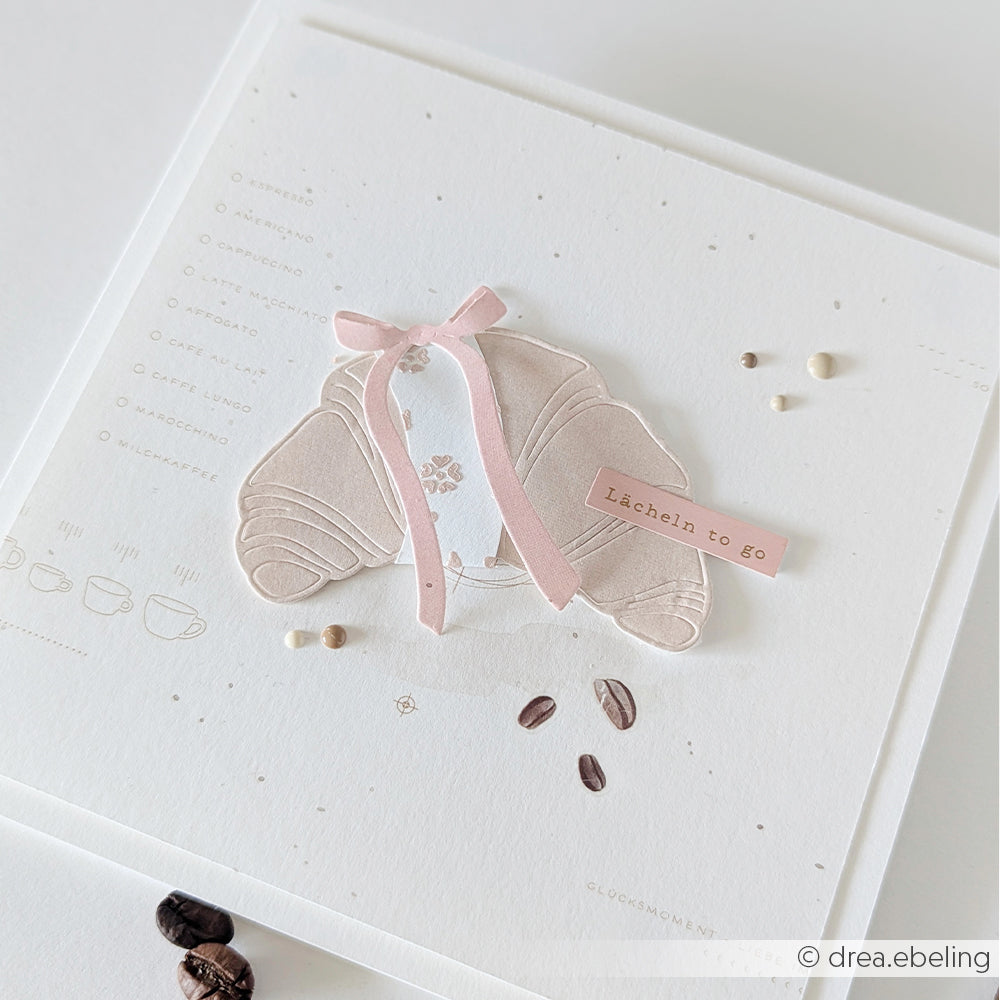 Eine handgefertigte Karte von Alexandra Renke aus Designpapier mit einer Croissant-Illustration, einer rosa Schleife und einem Etikett mit der Aufschrift „Lächeln to go“.