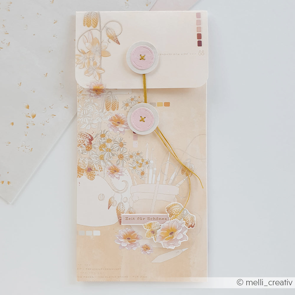Dekorativer beiger Umschlag von Alexandra Renke, gefertigt aus Designpapier mit rosa Knopfverschlüssen, Blumenillustrationen, die von der „Florale Wollkomposition Warmapricot“ inspiriert sind, und einer goldenen Kordel auf weißem Hintergrund – perfekt für Papierprojekte.