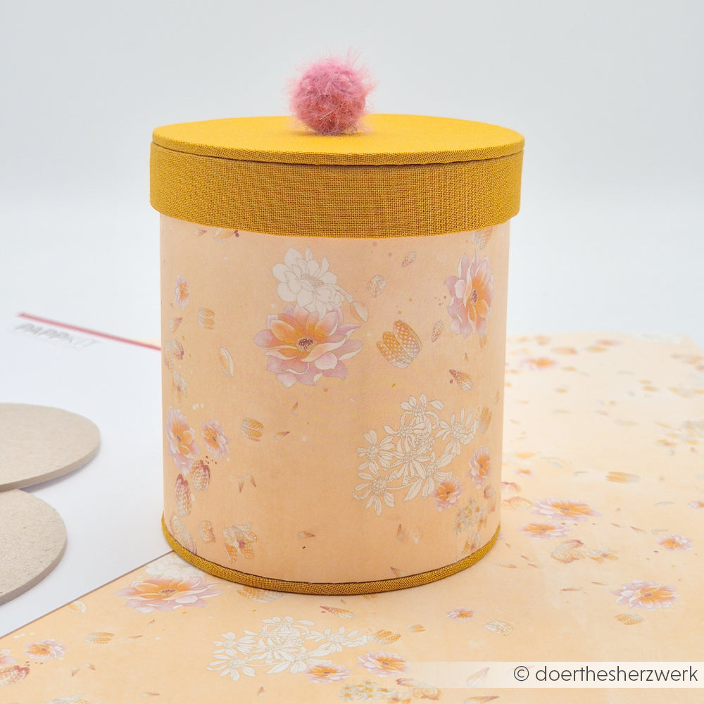 Eine zylindrische Blumenschachtel von Alexandra Renke mit gelbem Deckel und rosa Pompon, gefertigt aus Designpapier, steht auf einer passenden Unterlage – ein ideales Highlight für Papierprojekte oder jede beliebige florale Blütenkomposition.