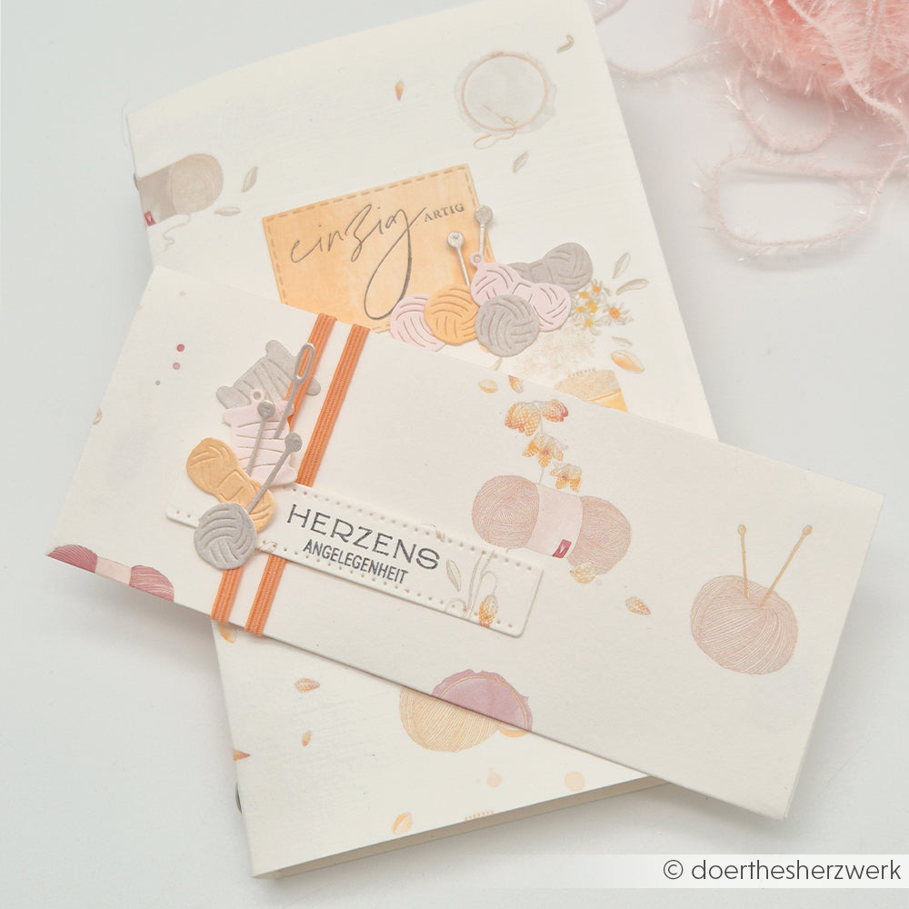 Eine Grußkarte zum Thema Stricken sowie ein passender Umschlag von Alexandra Renke, gefertigt aus Designpapier und mit deutschem Text, präsentiert auf weißem Hintergrund.