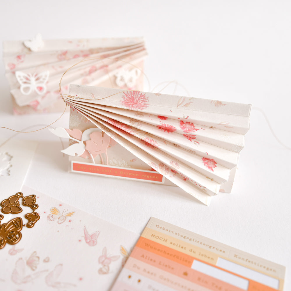 Eine aus Papier gefaltete Karte im Ziehharmonika-Stil mit zarten Schmetterlingsausschnitten, einer Schnur und pastellfarbenem Briefpapier von Alexandra Renke, präsentiert auf einem weißen Untergrund – perfekt für kreative Papierprojekte mit Designpapier und zarten Blumenmotiven.