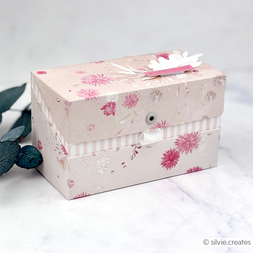 Eine blumig-rosa-weiße Dekorationsschachtel mit einem Anhänger, gefertigt aus elegantem Alexandra-Renke-Designpapier, präsentiert auf einer hellen Marmorfläche neben Eukalyptusblättern.