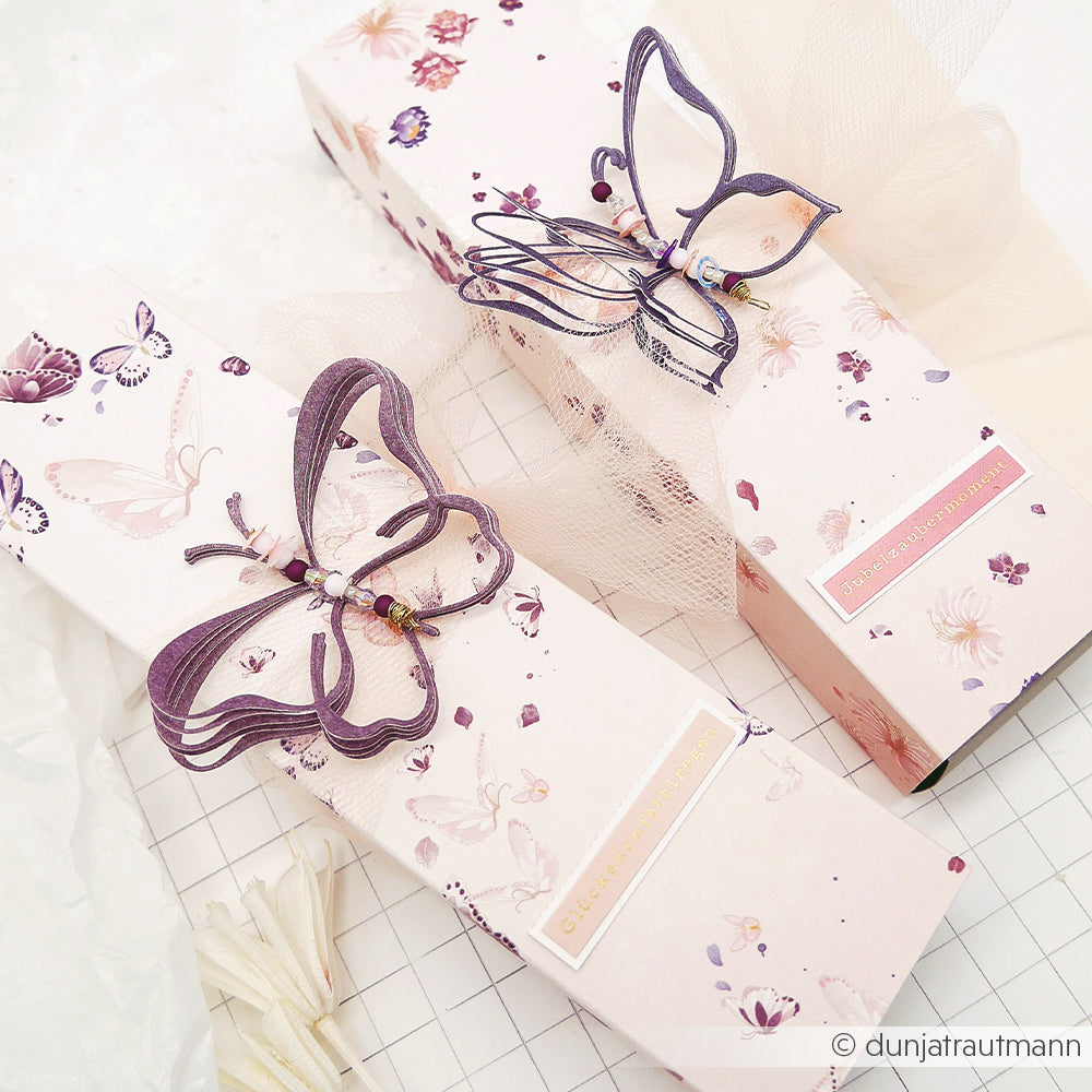 Zwei rosa Geschenkboxen mit Blumenmotiv von Alexandra Renke, gefertigt aus Designpapier, zeigen eine florale Schmetterlingskomposition mit zarten violetten Schmetterlingsverzierungen und Tüllschleifen vor einem Gitterhintergrund – perfekt für inspirierende Papierprojekte.