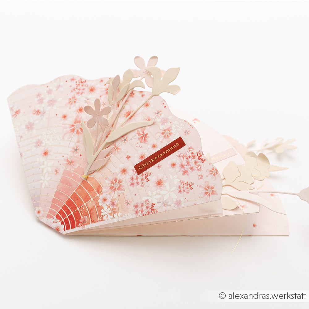 Eine rosafarbene Blumen-Pop-up-Karte von Alexandra Renke aus Designpapier mit ausgeschnittenen Blumen und "Glücksmoment" auf der Vorderseite. Ideal für Papierprojekte oder als Florale Foto Figurine Blassrosa als Andenken.