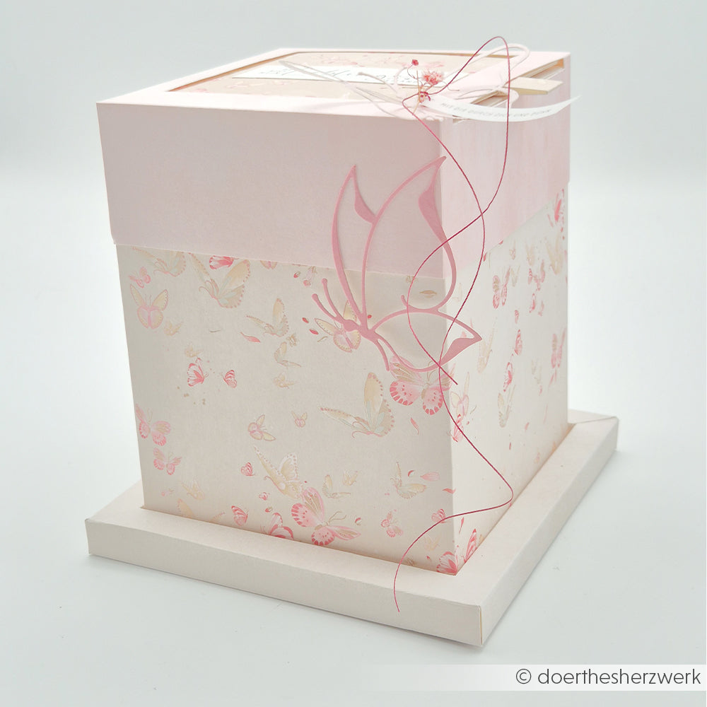 Quadratische rosa-weiße Geschenkschachtel von Alexandra Renke mit floralem Schmetterlingsmotiv und zartem Band, hergestellt aus hochwertigem Designpapier, auf weißem Hintergrund.