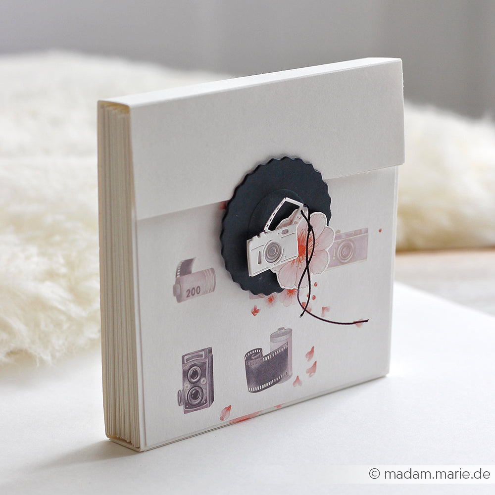 Ein weißer Umschlag mit Kamera- und Filmmotiven, gefertigt aus Designpapier und verziert mit einem dezenten floralen Fotomuster, verschlossen mit schwarzem, gewelltem Papier und einer Schnur – perfekt für einzigartige Papierprojekte.