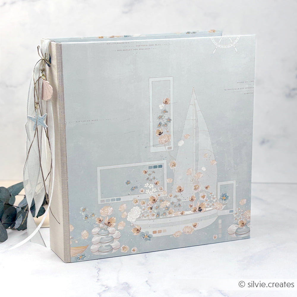 Alexandra Renke: hellblaues Scrapbook-Album mit Blumen- und Segelbootmotiven, gefertigt aus Designpapier und am Buchrücken mit Bändern sowie kleinen Anhängern verziert.