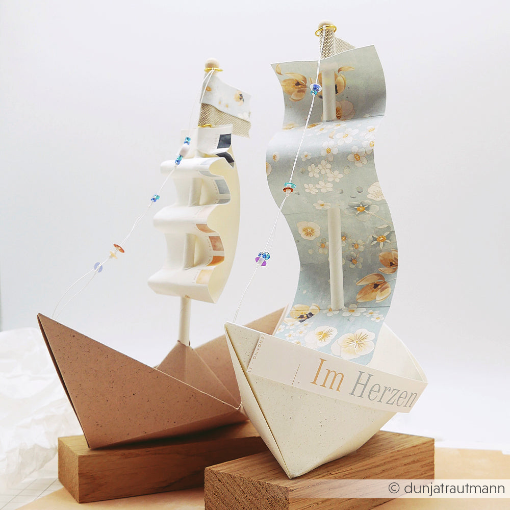 Zwei dekorative Papiersegelboote mit gemusterten Segeln aus Alexandra Renke Designpapier, ausgestellt auf Holzständern vor einem hellen Hintergrund.
