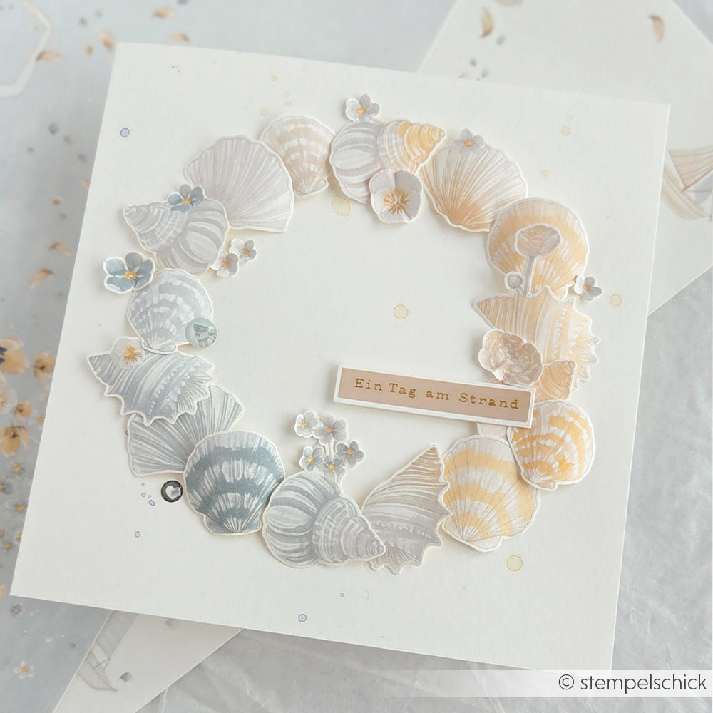 Quadratische Karte von Alexandra Renke mit einem pastellfarbenen Muschelkranz rund um den Schriftzug „Ein Tag am Strand“. Ideal für Papierprojekte oder als einzigartiges Designpapier.