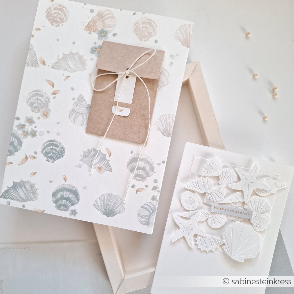 Zwei Karten mit Muschelmotiven, gestaltet aus hochwertigem Designpapier von Alexandra Renke: Die eine ist mit einem Kraftumschlag und einer Schnur verziert, während die andere ein mehrschichtiges weißes Muschelmotiv zeigt, das von einem floralen Muschelrapport inspiriert ist.