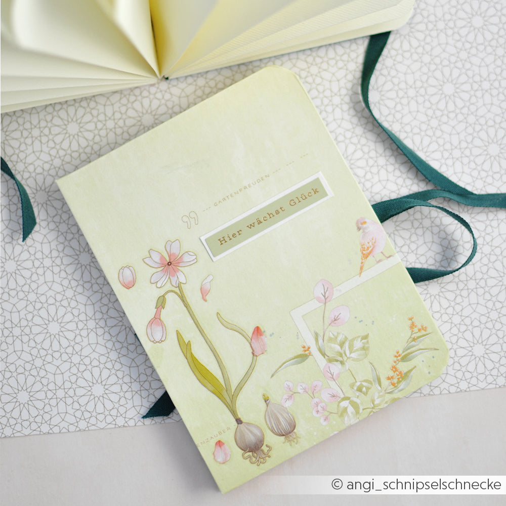Notizbuch mit pastellfarbenen Blumenmotiven von Alexandra Renke und dem deutschen Schriftzug „Hier wächst Glück“ auf gemustertem Hintergrund – ideal für Papierprojekte oder eine florale Gartenkomposition im Gelbgrün-Thema.