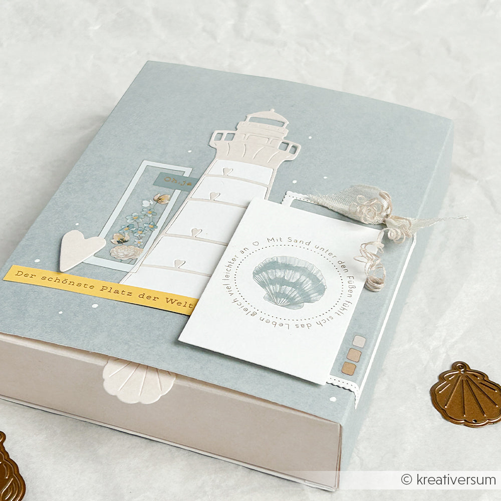 Blaugraue Geschenkbox mit einer Leuchtturm-Illustration und einer Karte mit Muschelmotiv, gefertigt aus Designpapier von Alexandra Renke und verziert mit Bindfaden, einem Herz und einer Karte mit Blumenmotiv.