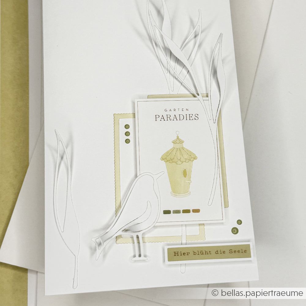 Weiße Karte von Alexandra Renke aus hochwertigem Designpapier, mit geprägten Blättern, Vogel- und Vogelhaus-Illustration sowie dem Text "Garten Paradies" - ideal für kreative Papierarbeiten.