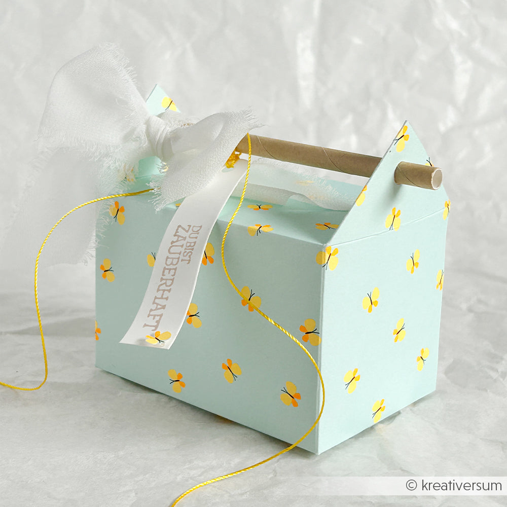 Eine hellblaue Geschenkbox aus Neon-Papier, verziert mit gelben Blumen, einer weißen Schleife und einem Anhänger, präsentiert vor einem weißen Hintergrund.