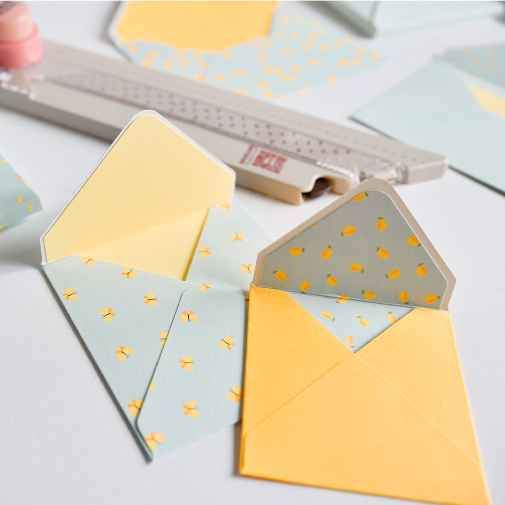 Auf einem weißen Tisch liegen zwei kleine pastellfarbene Umschläge, einer offen und einer geschlossen, mit gelben und zitronengelben Mustern auf Designpapier.
