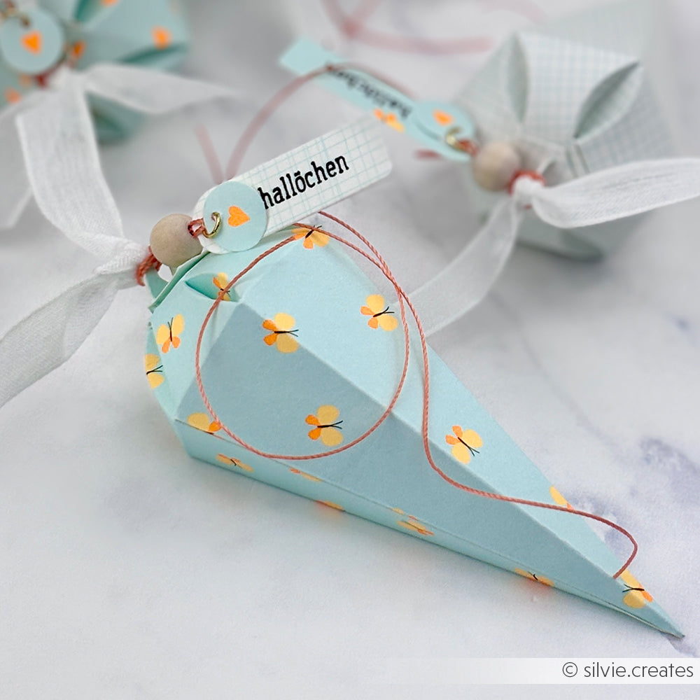 Hellblaue, kegelförmige Geschenkbox mit gelben Blumen, weißem Band, einer Holzperle und einem Anhänger mit der Aufschrift „hallöchen“. Als verspielte Note sind zarte Schmetterlinge aus leuchtendem Neonpapier darauf angebracht.