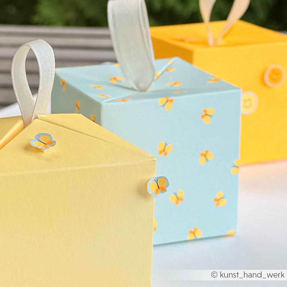 Drei pastellfarbene Geschenkboxen – zwei gelbe und eine blaue mit gelben Blumen –, jeweils mit einer Schleife oben – perfekt für Ihre „Sommer Special Schmetterlinge Papierprojekte“ oder als reizvolle Ergänzung zu jedem Designpapier-Projekt.