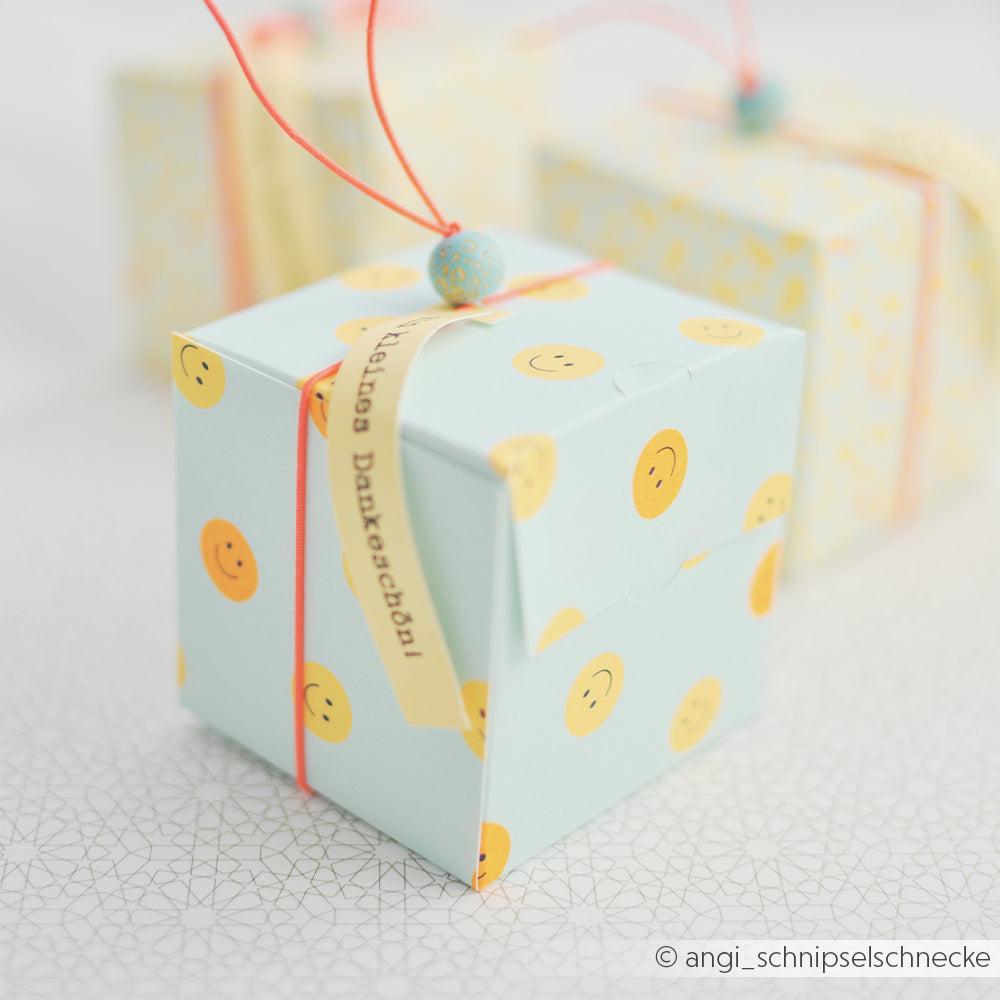 Hellblaue Geschenkbox mit „Summer Special“-Smileys und gelben Smileys, verziert mit einer orangefarbenen Schnur und einer blauen Perle, gefertigt aus hochwertigem Designpapier von Papierprojekte auf weißem Untergrund.