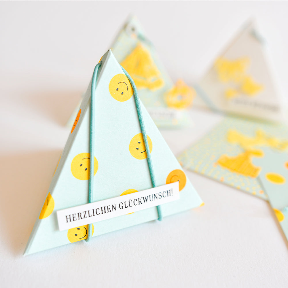 Dreieckige Geschenkbox mit gelben Smileys und einem Aufkleber mit der Aufschrift „HERZLICHEN GLÜCKWUNSCH!!“ – perfekt für Papierprojekte und aus farbenfrohem Designpapier gefertigt, das für eine fröhliche Note sorgt.