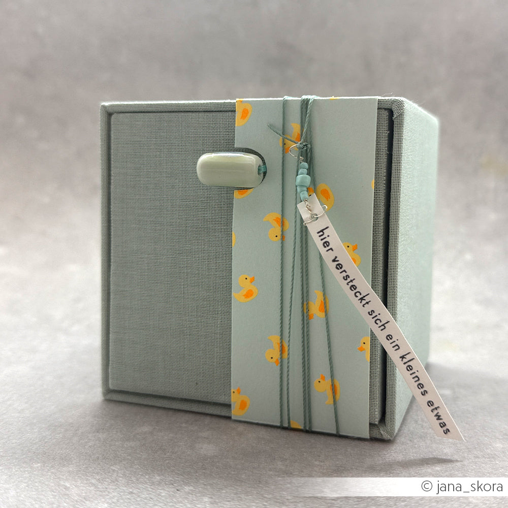 Eine kleine graue Schachtel mit gelben Entchenmotiven aus hochwertigem Designpapier ist mit einem Knopf und einem Anhänger mit der Aufschrift „hier versteckt sich ein kleines etwas“ versehen. Dieses entzückende „Sommer-Special-Entchen“-Accessoire verleiht jeder Neon-Papier-Kollektion eine verspielte Note.