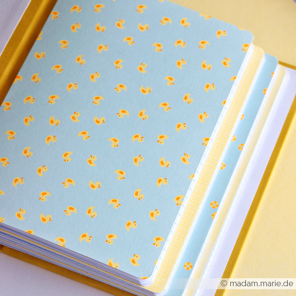 Eine Nahaufnahme von Notizbüchern mit blauen Einbänden, die mit einem Muster aus kleinen gelben Gummienten verziert sind – ideal für Papierprojekte oder als verspielte Akzente auf Designpapier.