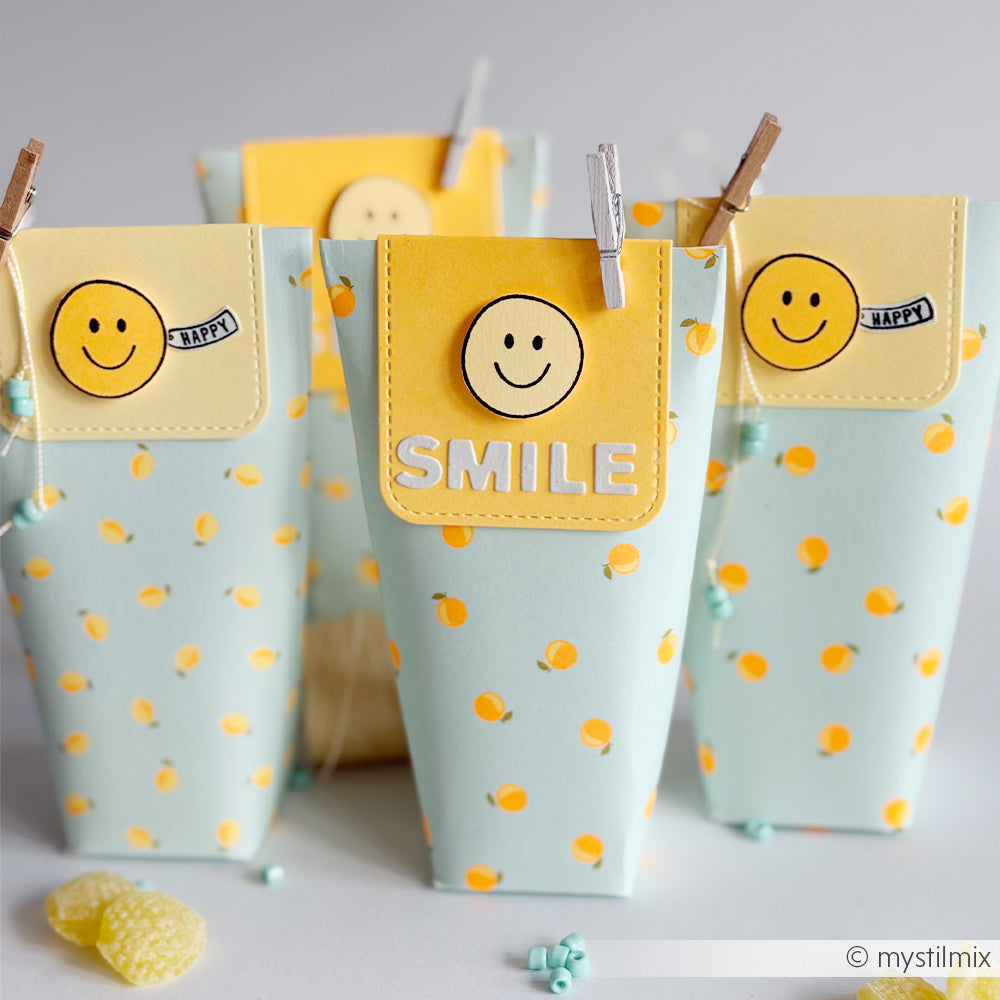 Vier pastellfarbene Geschenktüten mit gelben Smileys und der Aufschrift „SMILE“, gefertigt aus hochwertigem Designpapier und verziert mit Polka-Dots und Clips – perfekt für Ihre Papierprojekte oder als Teil einer Feier zum Thema „Sommer-Special Orangen“.