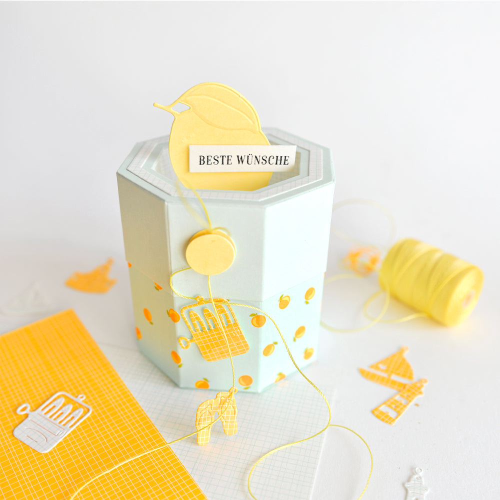 Achteckige Geschenkbox aus Designpapier mit gelben Punkten, mit einem Vogelausschnitt, einer Karte mit der Aufschrift „Beste Wünsche“ und einem gelben Faden daneben – perfekt für kreative Papierprojekte.