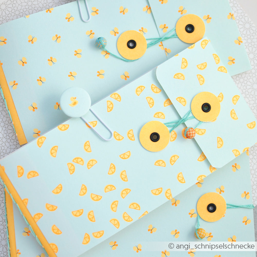 Hellblaue Umschläge mit gelben Zitronenscheiben-Motiven aus der Sommer-Special-Kollektion „Zitronenspalten Designpapier“, versehen mit Schnurverschlüssen und so angeordnet, dass sie sich überlappen – perfekt, um Ihren Papierprojekten eine frische Note zu verleihen.