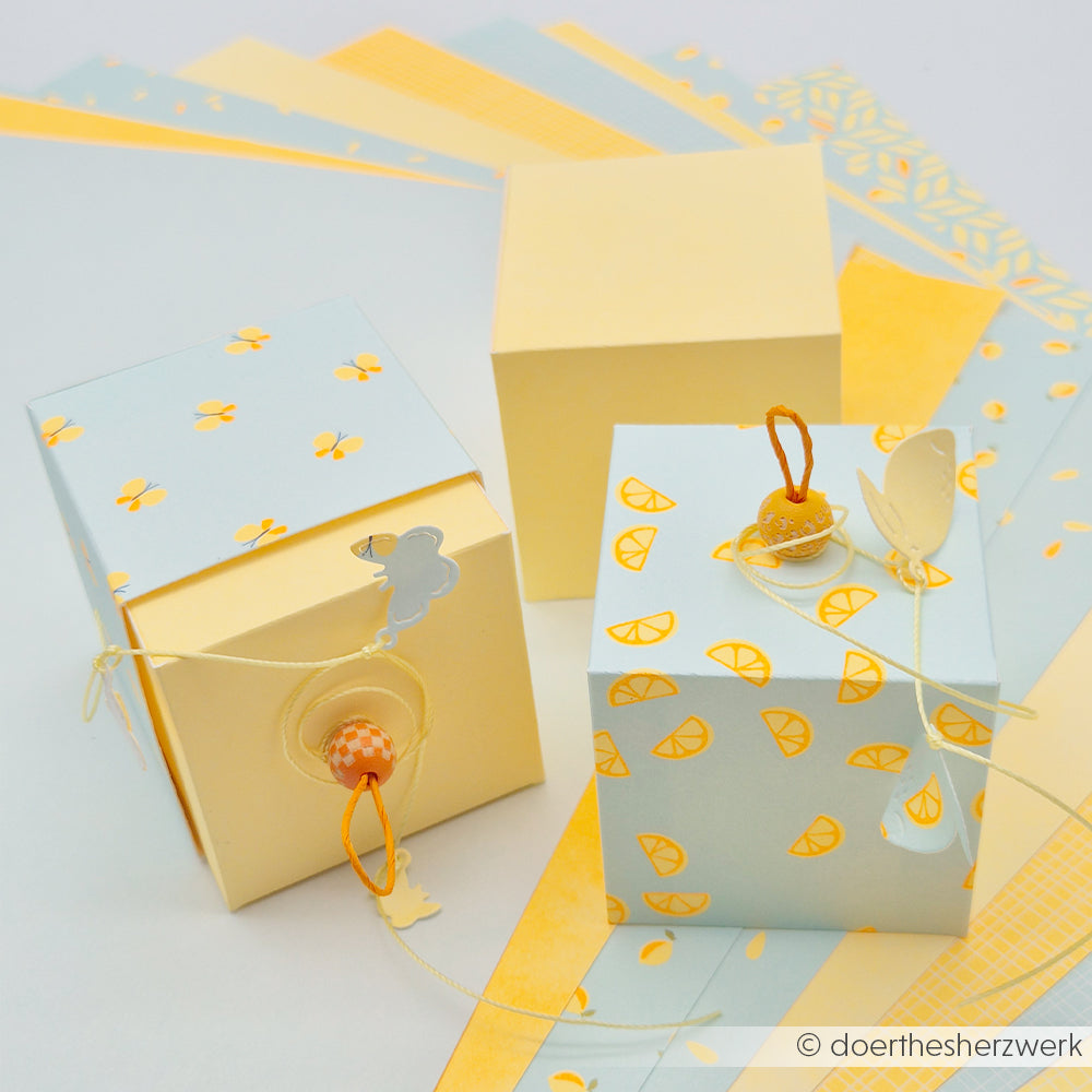 Auf einem gelb-weißen Hintergrund sind zwei pastellfarbene Geschenkboxen mit gelben Mustern und Papierverzierungen zu sehen, die aus Neonpapier und Designpapier gefertigt sind.