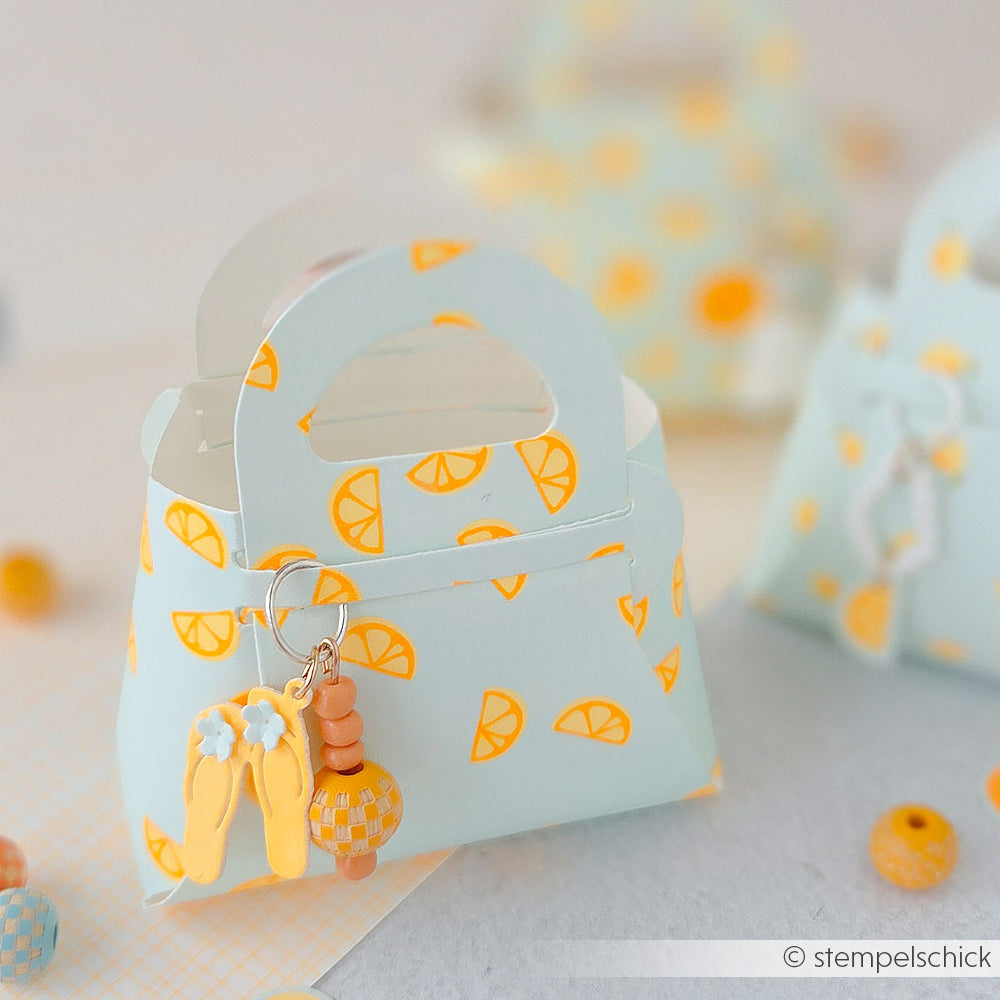 Hellblaue Designpapier-Tasche mit einem fröhlichen gelb-orangefarbenen Zitronenscheiben-Aufdruck – perfekt für Ihre „Sommer Special Zitronenspalten“-Papierprojekte – komplett mit einem Flip-Flop-Schlüsselanhänger vor einem sanften Pastellhintergrund.