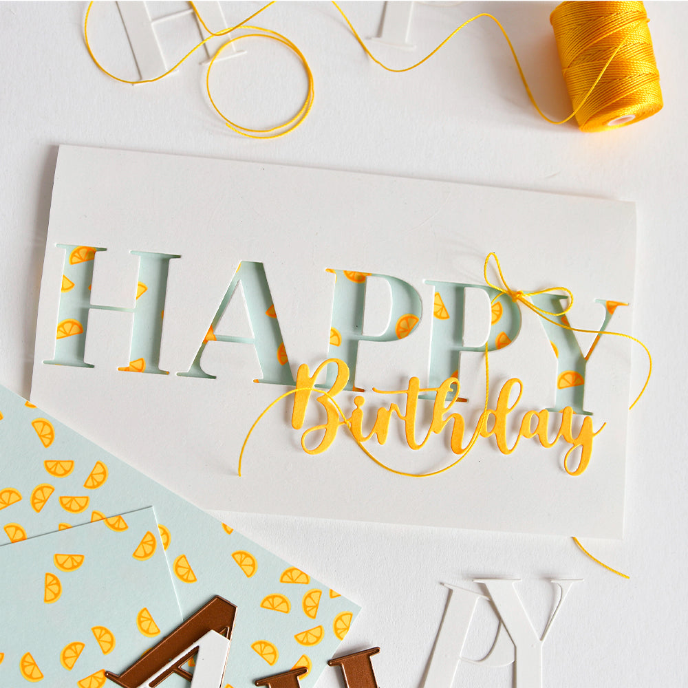 Weiße Karte mit der Aufschrift „HAPPY Birthday“, Orangenscheiben, gelbem Faden und Bastelmaterial auf weißem Untergrund – ideal für Papierprojekte oder um mit Designpapier und Neonpapier einen Farbtupfer zu setzen.
