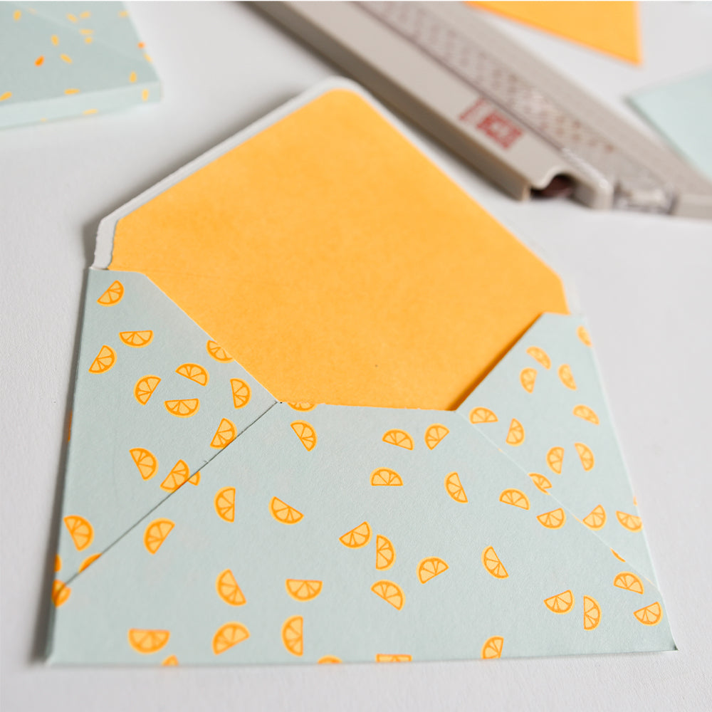 Offener Umschlag mit einem Muster aus orangefarbenen Zitrusscheiben, gefertigt aus leuchtendem Neon-Papier, mit einer schlichten orangefarbenen Karte auf weißem Hintergrund.