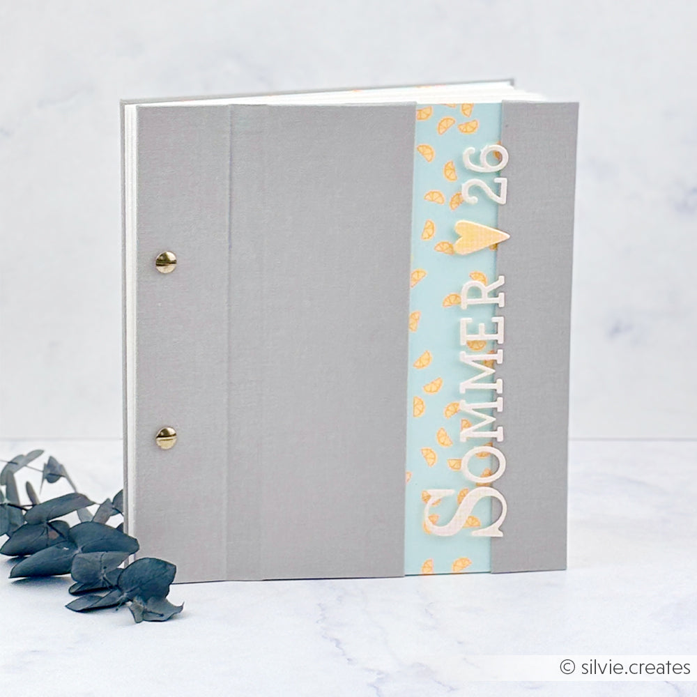 Graues Scrapbook-Album mit der Aufschrift „SOMMER ♥ 26“ auf einem blauen Streifen, gefertigt aus hochwertigem Designpapier, daneben einige grüne Eukalyptusblätter.